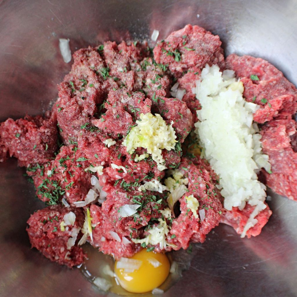 Raw ground beef, chopped onion, minced garlic, eggs, and seasonings in a bowl
