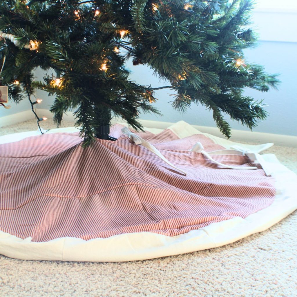 Red pinstripe tree skirt with white ruffle and ties underneath a tree with Christmas lights 