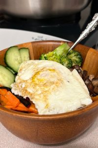 bamboo bowl filled with cucumbers, meat, carrots, rice, broccoli, and topped with a fried egg. Bosch mixer in the background