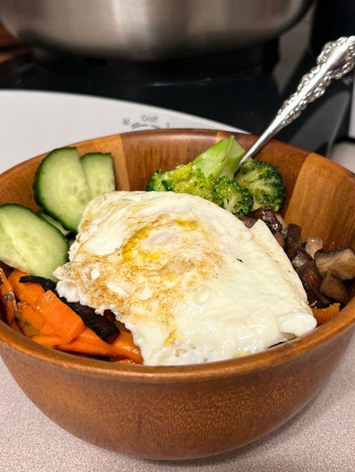 bamboo bowl filled with cucumbers, meat, carrots, rice, broccoli, and topped with a fried egg. Bosch mixer in the background